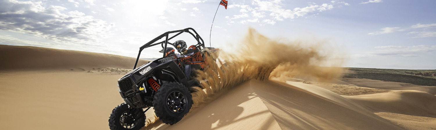 Two people riding in a Polaris® RZR® ATV on dunes while kicking up a cloud of sand on a sunny day.